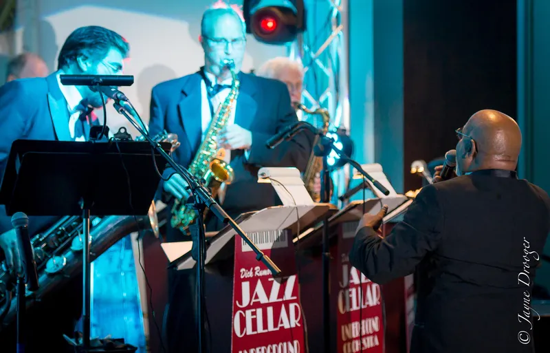 Tuxedoed saxophonists play behind red “Jazz Cellar” music stands as a bald vocalist in black faces the band