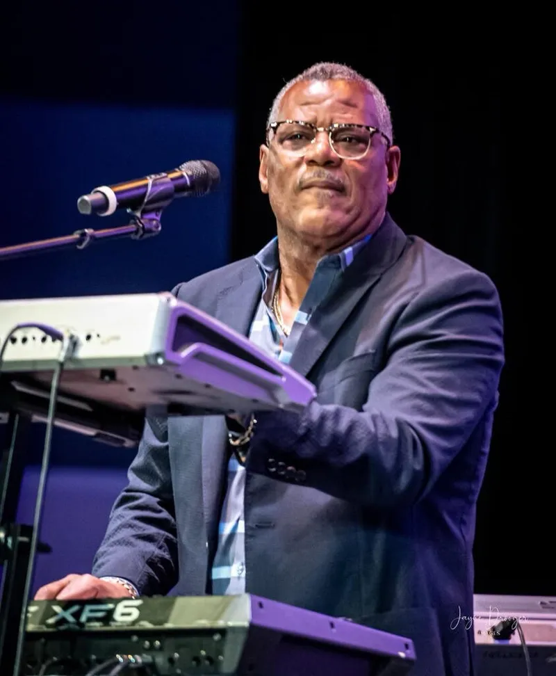 Shawn Brown in glasses and a navy blazer at his stacked keyboards beside a microphone on a dark stage