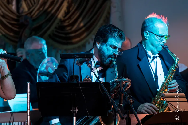 Two tuxedoed saxophonists play under teal stage lights beside music stands, with ornate draped curtains behind