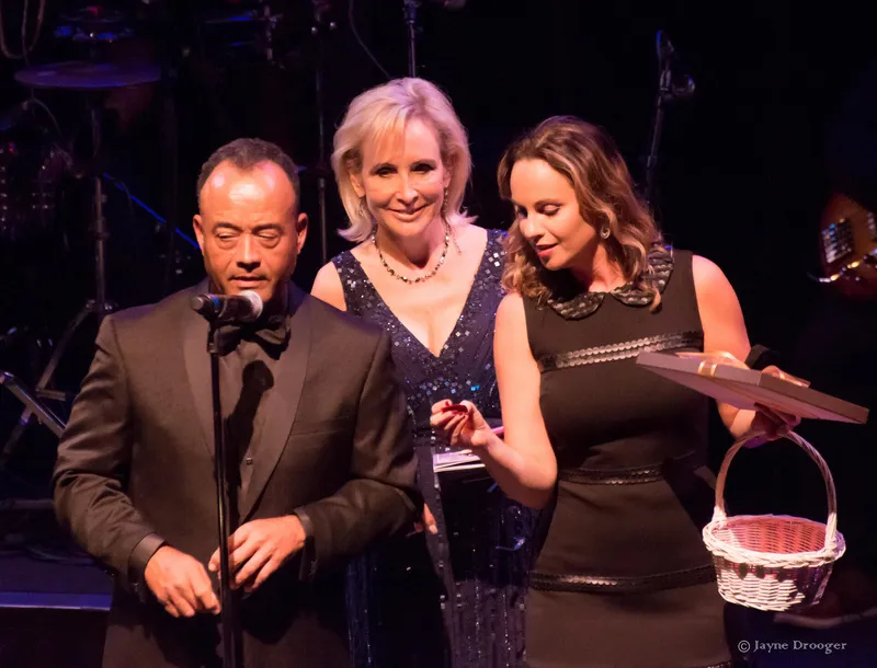 A tuxedoed man at a microphone with a woman in a sequined gown and another holding a gift and basket