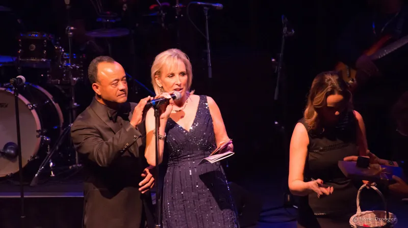 A woman in a sequined navy gown speaks into a microphone as a tuxedoed man adjusts it on stage