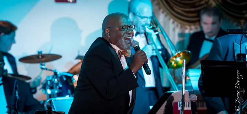 A smiling bald vocalist with a gray beard, glasses and a patterned bow tie sings with the big band