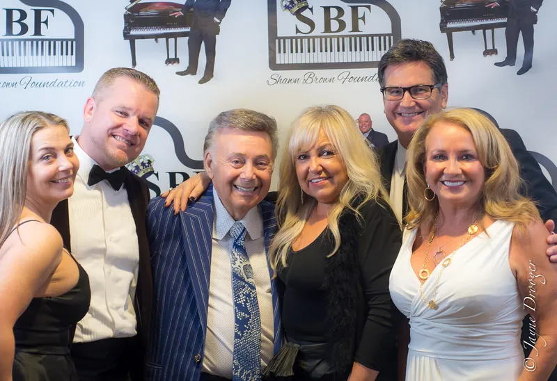 Six smiling guests in evening wear pose together in front of a Shawn Brown Foundation logo backdrop