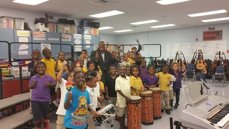 A man in a suit gives a thumbs-up among smiling schoolchildren holding drums and percussion in a school music room