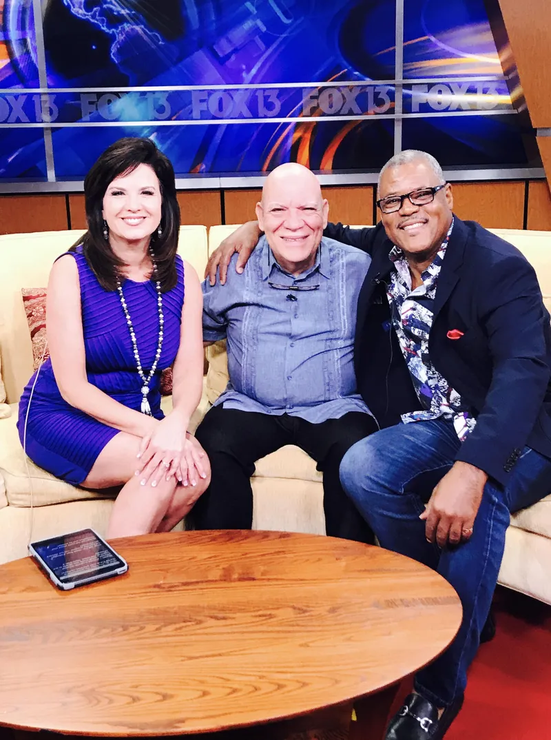Shawn Brown sits on a FOX 13 studio couch with a woman in a purple dress and a smiling bald man