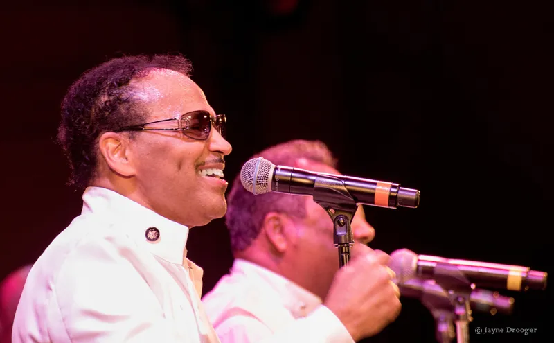 A smiling singer in tinted sunglasses and a white jacket performs at a microphone beside another singer in white
