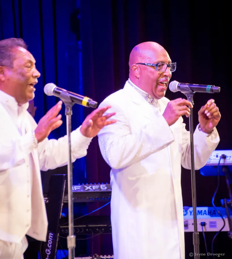 Two vocalists in white suits sing and gesture at microphones, one bald with glasses, keyboards behind them