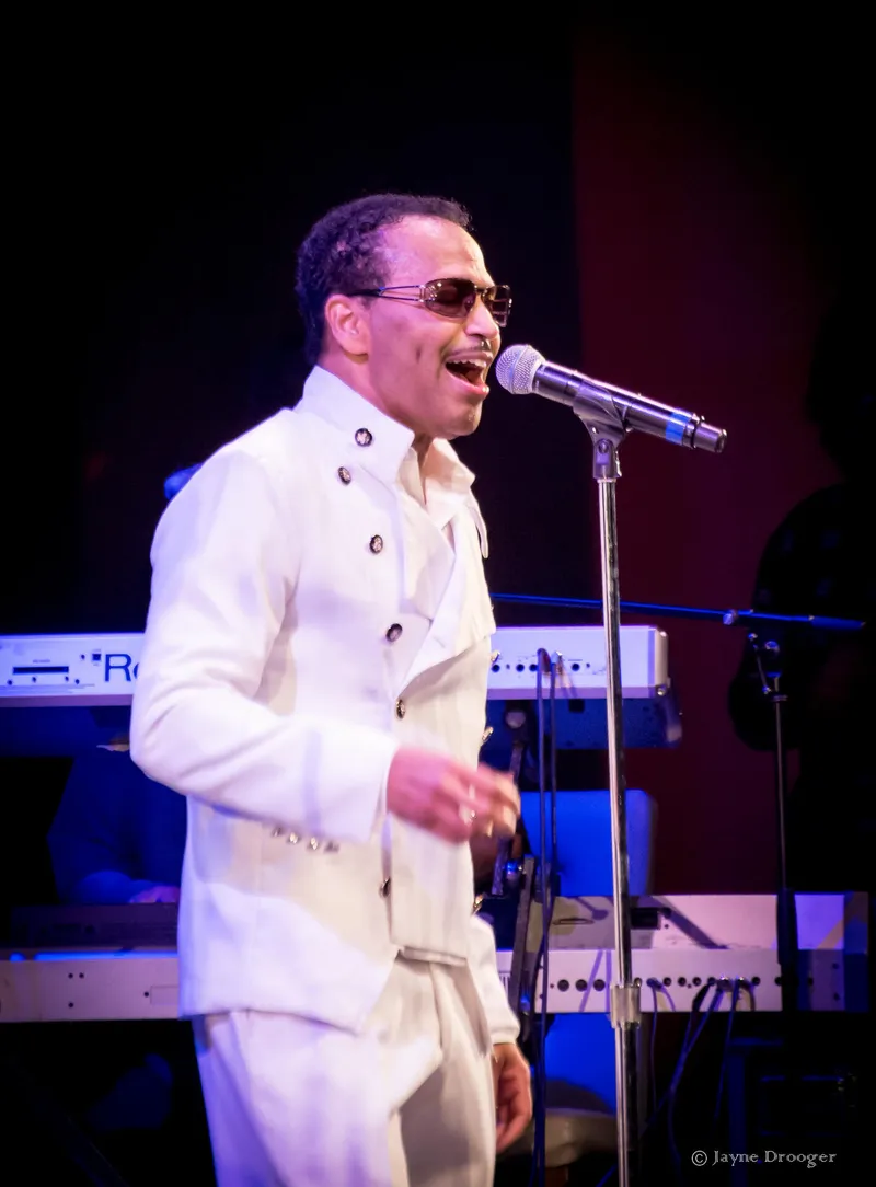 A singer in a white double-breasted jacket and tinted sunglasses sings at a microphone stand before keyboards