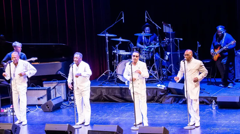Wide stage view of four vocalists in white suits at microphones, backed by drums, bass, guitar and a grand piano