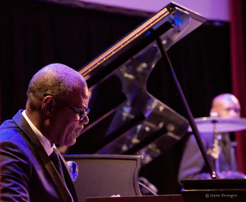 Shawn Brown, in glasses and a dark suit, plays in profile beside a grand piano with its lid raised