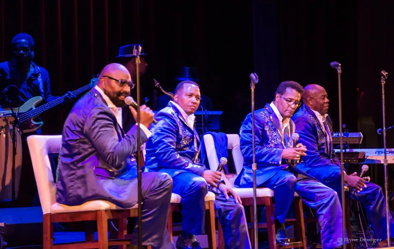 Four vocalists in shiny blue embroidered suits sit on white chairs as a bearded singer speaks, band behind them