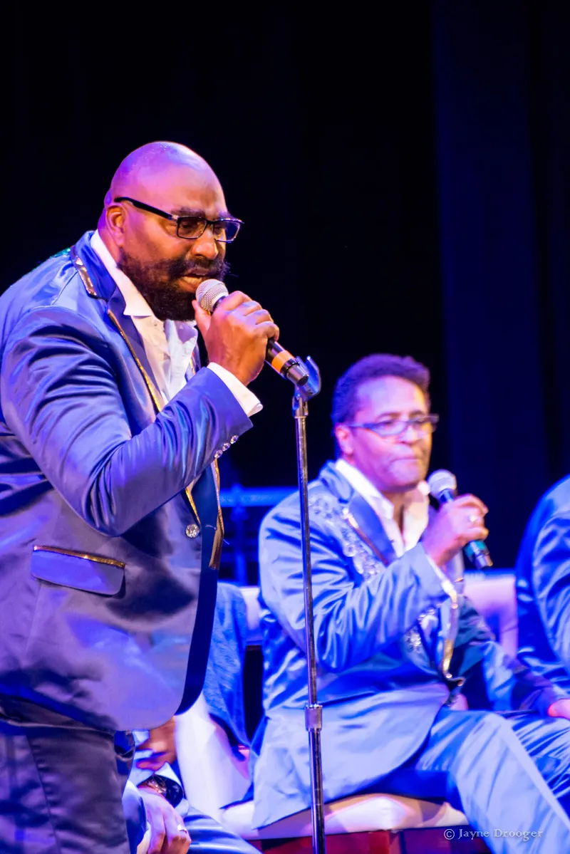 A bearded, bald singer in glasses and a shiny blue jacket sings into a microphone, a seated bandmate behind