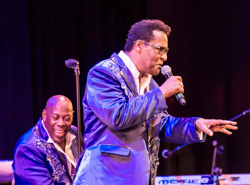 A singer in glasses and an embroidered blue satin jacket sings and gestures as a smiling bandmate looks on