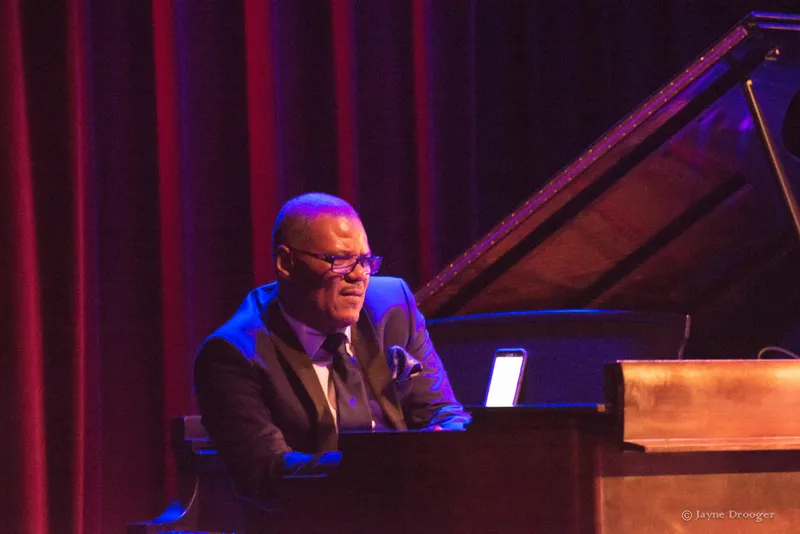 Shawn Brown in glasses and a dark suit plays a wooden organ, with red curtains and a grand piano behind