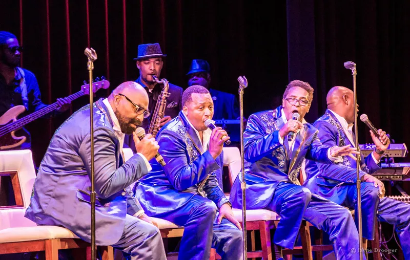 Four vocalists in blue satin suits sing seated at microphones, with a saxophonist in a fedora and a bassist behind
